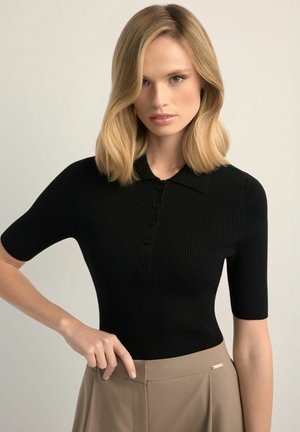 Blondynka o włosach do ramion, ubrana w czarny prążkowany top z krótkim rękawem i beżowe spodnie z wysokim stanem, pozująca z jedną ręką na biodrze.