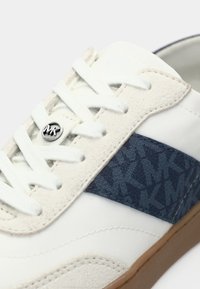 Baskets blanches avec une tige en daim et cuir, présentant un panneau latéral bleu à motif, des lacets blancs, et un accent de logo argenté circulaire.