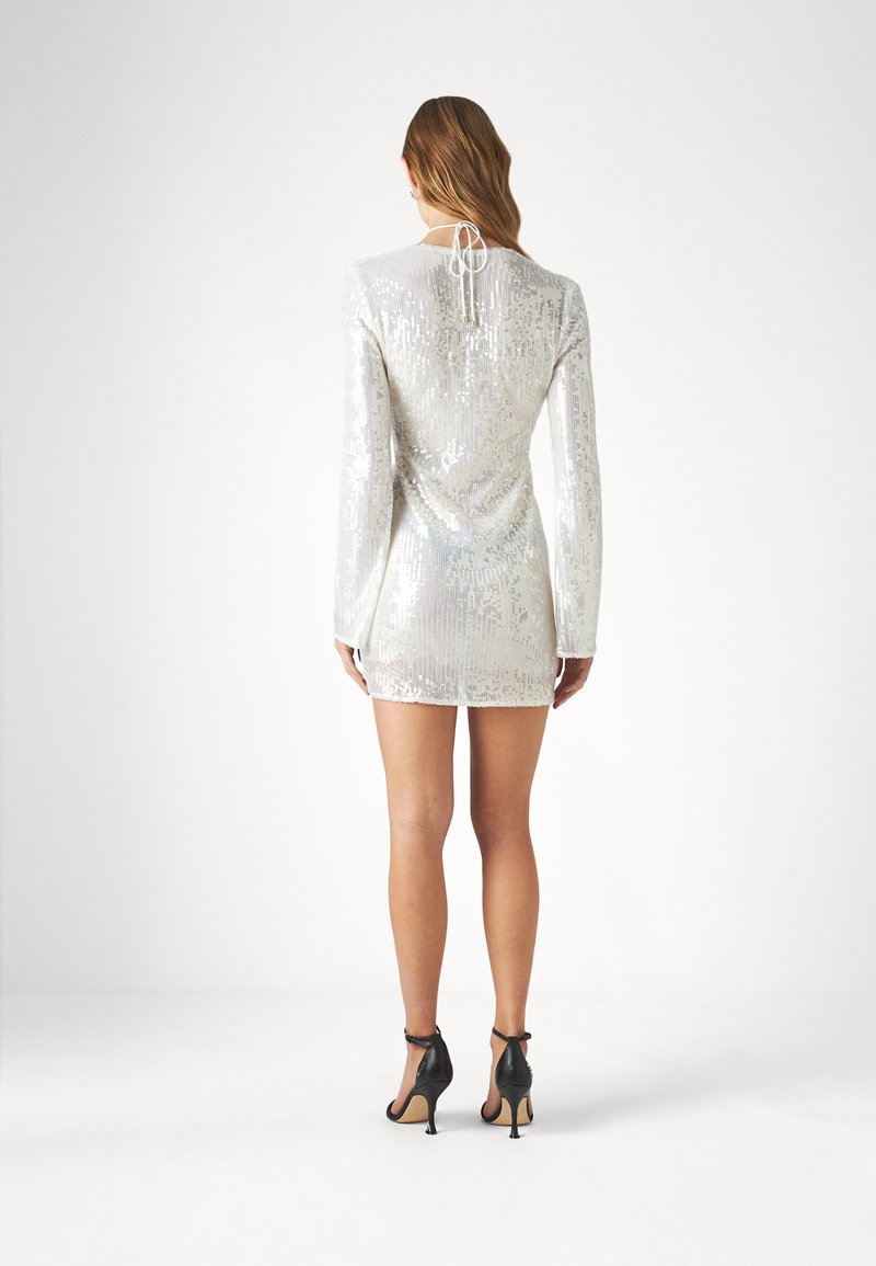 Srebrna sukienka z cekinami, z długimi rękawami, krótkim dołem i wiązaniem przy szyi. Modelka nosi czarne szpilki.