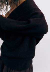 Persoon draagt een oversized donker gebreide trui met een diepe V-hals, los krullend haar gedeeltelijk zichtbaar, tegen een effen lichte achtergrond.