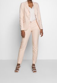 Costume rose clair sur mesure avec un blazer à simple boutonnage et un pantalon slim, associé à un haut blanc et des sandales blanches à lanières.