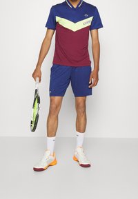 Lacoste tennisoutfit har en vinröd och blå färgblockerad polo med gröna detaljer, tillsammans med blå shorts och vita-oranga skor.