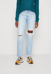 Modré džíny se středním praním a roztrženými koleny, kombinované s teal pleteným svetr a teniskami s béžovými akcenty. Pohodlný střih s třepenými okraji.