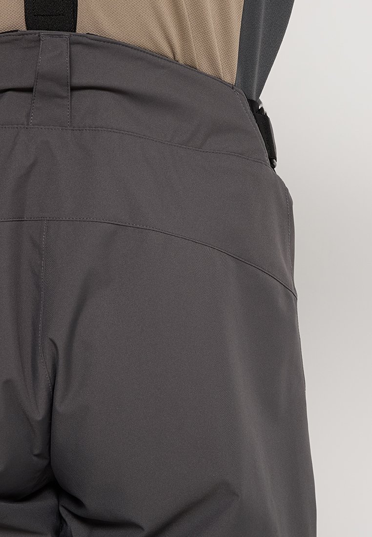Szare wodoodporne spodnie z wzmocnionymi szwami noszone na beżowej koszuli, z regulowanymi czarnymi szelkami po bokach.