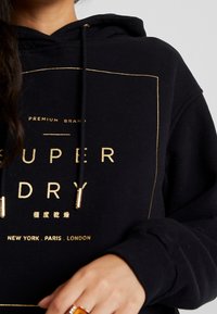 Kvinna som bär en svart Superdry-huvtröja med guldtext och dragsnören, som visar en del av hennes hand med en ring.