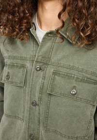 Chaqueta verde oliva de algodón con un acabado texturizado, que presenta dos bolsillos en el pecho, botones plateados y un cuello.
