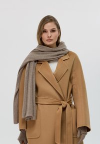 Cappotto di lana color tan con vita alta, tasche a soffietto e ampi rever, abbinato a una sciarpa di cashmere color marrone chiaro drappeggiata intorno al collo.