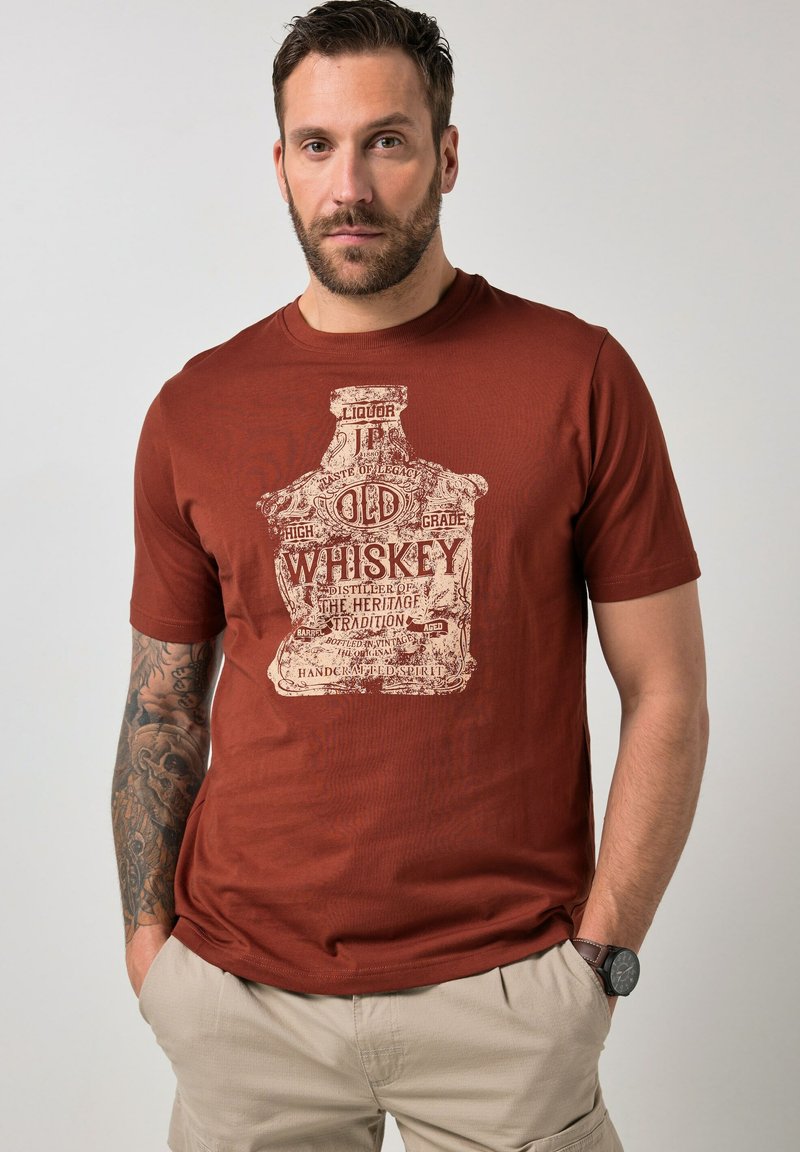 Uomo con barba e tatuaggio che indossa una maglietta color ruggine con grafica di bottiglia di whisky vintage e pantaloni beige, in piedi con le mani in tasca.