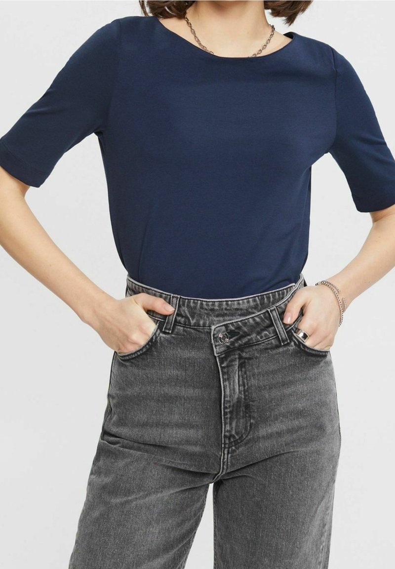 Personne portant un haut bleu marine à manches courtes rentré dans un jean noir délavé taille haute, les mains dans les poches avant.