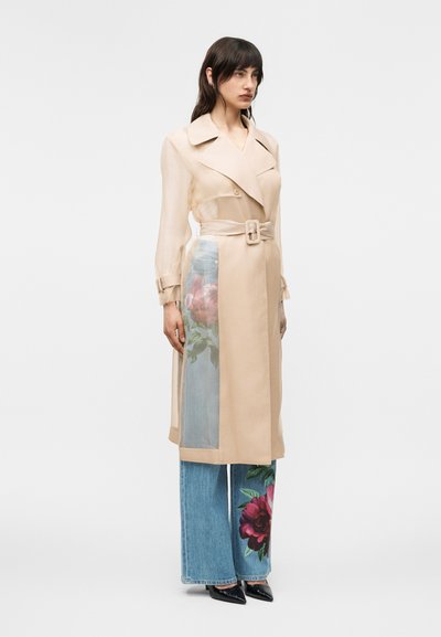 Femme portant un trench-coat beige ceinturé avec des panneaux transparents, par-dessus un jean bleu orné de grandes impressions florales et des chaussures noires à bouts pointus.