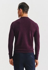 Mężczyzna ubrany w dopasowany, ciemnofioletowy sweter z długim rękawem oraz ciemnogranatowe spodnie, stojący tyłem na jednolitym tle.