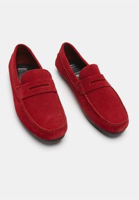 Crvene zeca loferice s ravnim đonom, s prošivenim dizajnom i uzdignutim prednjim dijelom. Unutrašnjost je crna, ističući stil slip-on.