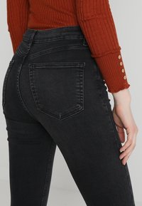 Vaqueros negros de tiro alto con piernas ajustadas y dos bolsillos traseros, hechos de denimb con una textura suave. Se combinan con un suéter naranja de canalé.