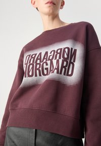 Sweat-shirt bordeaux avec une bande dégradée argentée comportant un texte inversé. Tissu doux, coupe ample, col et ourlet côtelés. Pantalon en cuir noir visible.