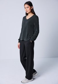 Dunkelgrünes Langarmoberteil mit V-Ausschnitt, kombiniert mit dunklen Nadelstreifen-Hosen und hellgrauen Sneakers, die eine lockere Passform und glatte Textur aufweisen.