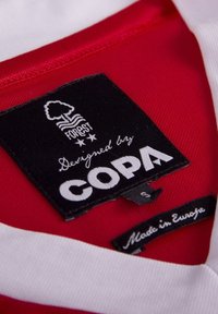 Rød og hvid bomuldsbeklædningsmærketag med en sort label med et trælogo, "Designet af COPA" og "Fremstillet i Europa" tekst. Størrelse: S.