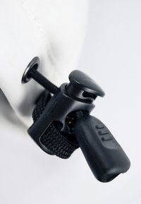 Un taquet en plastique noir et un verrou de cordon attachés à un tissu blanc, présentant une texture tressée, une forme ronde et une finition mate.