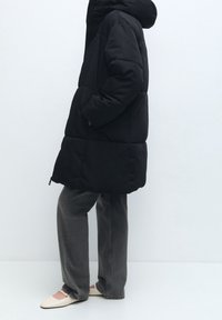 Černý prošívaný kabát s kapucí, se zipem, bočními kapsami a měkkým texturovaným povrchem, zkombinovaný se šedými kalhotami a světlými botami.