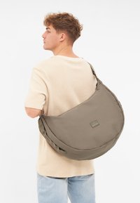 Borsa a spalla grigia realizzata in tessuto morbido, caratterizzata da una forma rotonda, una fascia larga e un piccolo logo sul davanti. Indossata a tracolla.