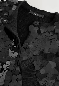Black cardigan featuring a textured knit and large black sequins. Two buttons at the neck and a smooth fabric interior are visible.