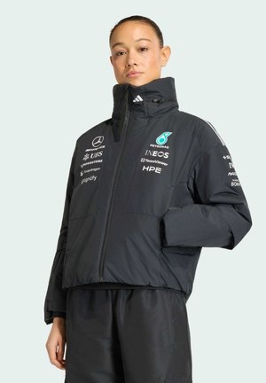 Femme portant une veste noire zippée avec les logos de l'équipe Mercedes AMG Petronas F1, debout devant un fond clair uni, mains dans les poches.