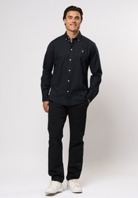 Schwarzes Hemd mit Knopfverschluss und weißen Knöpfen, taillierte Passform. Getragen mit schwarzen Hosen und weißen Sneakers. Verziert mit kleinem gestickten Logo auf der Brust.