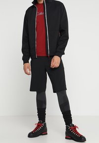 Veste noire zippée portée sur un t-shirt rouge, associée à un short noir et des leggings noirs à motifs. Bottes noires avec accents rouges portées.