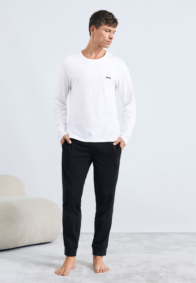 Valkoinen pitkähiha shirt, jossa on pieni musta logo rinnassa, yhdistetty mustiin kapeneviin housuihin. Minimalistinen muotoilu, pehmeä kangas, rento istuvuus.