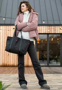 Černá taška přes rameno s dlouhými pop Ručkami, texturovaná látka, ohnuté okraje, růžové logo. Modelka nosí světle růžovou péřovou bundu, bílý pletený svetr a tmavé džíny.