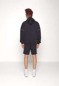 Felpa blu scuro con cappuccio e piping bianco, abbinata a pantaloni corti coordinati. Indossata con sneaker chiare, mostrando la vista posteriore.