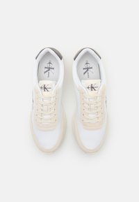 Sneakers Calvin Klein con tomaia in tela bianca, dettagli beige, punta rotonda, lacci piatti e linguetta posteriore grigia. Logo sulla linguetta.