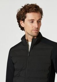Svart zip-up jacka med en vadderad framdel, släta ärmar och en uppstående krage. Texturerat tyg med en elegant, minimalistisk design.