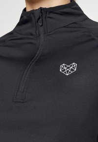 Černý sportovní top s zipem na čtvrt. Na hrudi je geometrické bílé srdíčko. Hladká, přiléhavá tkanina s minimálním počtem švů.