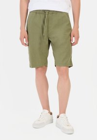 Olivengrønne linneshorts med elastisk talje og snor. Har en løs pasform og knælangt snit. Bæres med hvide sneakers.