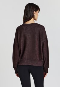 Maglione morbido e svasato di colore marrone scuro, realizzato in un tessuto textured, con orlo a costine e spalle scese. Indossato con leggings neri, evidenziando uno stile casual.