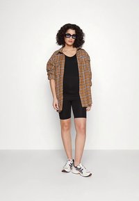 Chemise à carreaux marron oversize portée sur un débardeur noir et un short de cyclisme noir, avec des chaussures de course blanches et noires et des lunettes de soleil foncées.