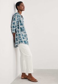 Blauwe en witte tuniek met patroon en driekwart mouwen, gecombineerd met witte broek en bruine sandaaltjes met bandjes. Glad stof, ontspannen pasvorm.