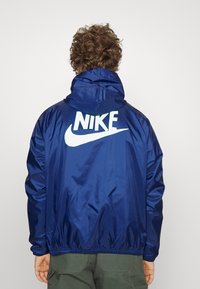 Marinblå, huvorjacka tillverkad av glänsande, lätt material. Har en stor vit "Nike"-logotyp och swoosh på ryggen. Elastiska ärmslut och nederkant.