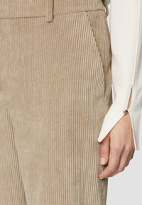 Beige corduroy broeken met verticale ribbelstructuur en een zijzak. Een witte trui met een split bij de pols en metalen knoopdetails is zichtbaar.
