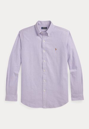 Jeune homme portant une chemise à boutons de couleur violet clair, un pantalon plissé taille haute beige avec une ceinture rayée, et des chaussures en cuir marron, debout les mains dans les poches.