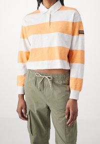 Gestreiftes orange-weißes langärmeliges Polohemd mit Kragen; weicher Stoff, cropped Design, mit einem Logo-Patch auf dem Ärmel, kombiniert mit olivgrünen Hosen.