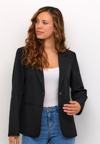 Černý blazer s přiléhavým střihem, jedním knoflíkem, klopami a dvěma předními kapsami, nošený přes bílý top a modré džíny.