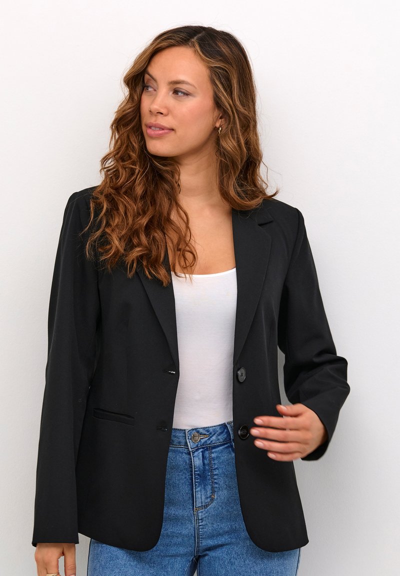 Černý blazer s přiléhavým střihem, jedním knoflíkem, klopami a dvěma předními kapsami, nošený přes bílý top a modré džíny.