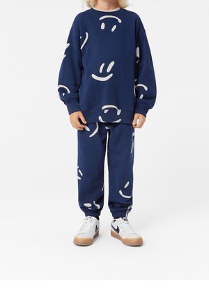 Ensemble de sweat-shirt et pantalon bleu marine avec des motifs de visages souriants blancs surdimensionnés, doté de poignets élastiques et de baskets dans des tons blancs et gris.