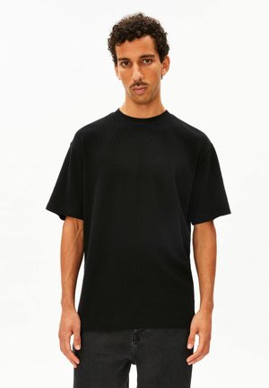 Man met kort krullend haar en snor, die een effen zwarte T-shirt met korte mouwen en een donkere broek draagt, staand tegen een witte achtergrond.