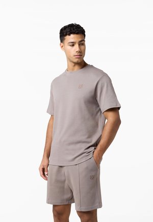 Mężczyzna ubrany w pasującą bluzkę z krótkim rękawem i szorty w kolorze taupe z subtelnym logo, stoi z jedną ręką w kieszeni, patrzy na bok.