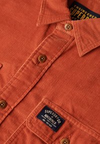 Rode corduroy overhemd met een button-down kraag, twee borstzakken en een label van stof met "Superdry Co"-branding.