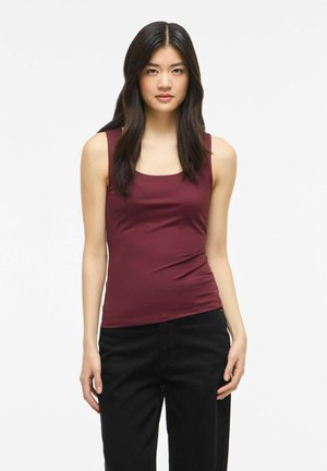 Jeune femme aux longs cheveux foncés portant un haut bordeaux sans manches et un pantalon noir, debout devant un fond blanc.