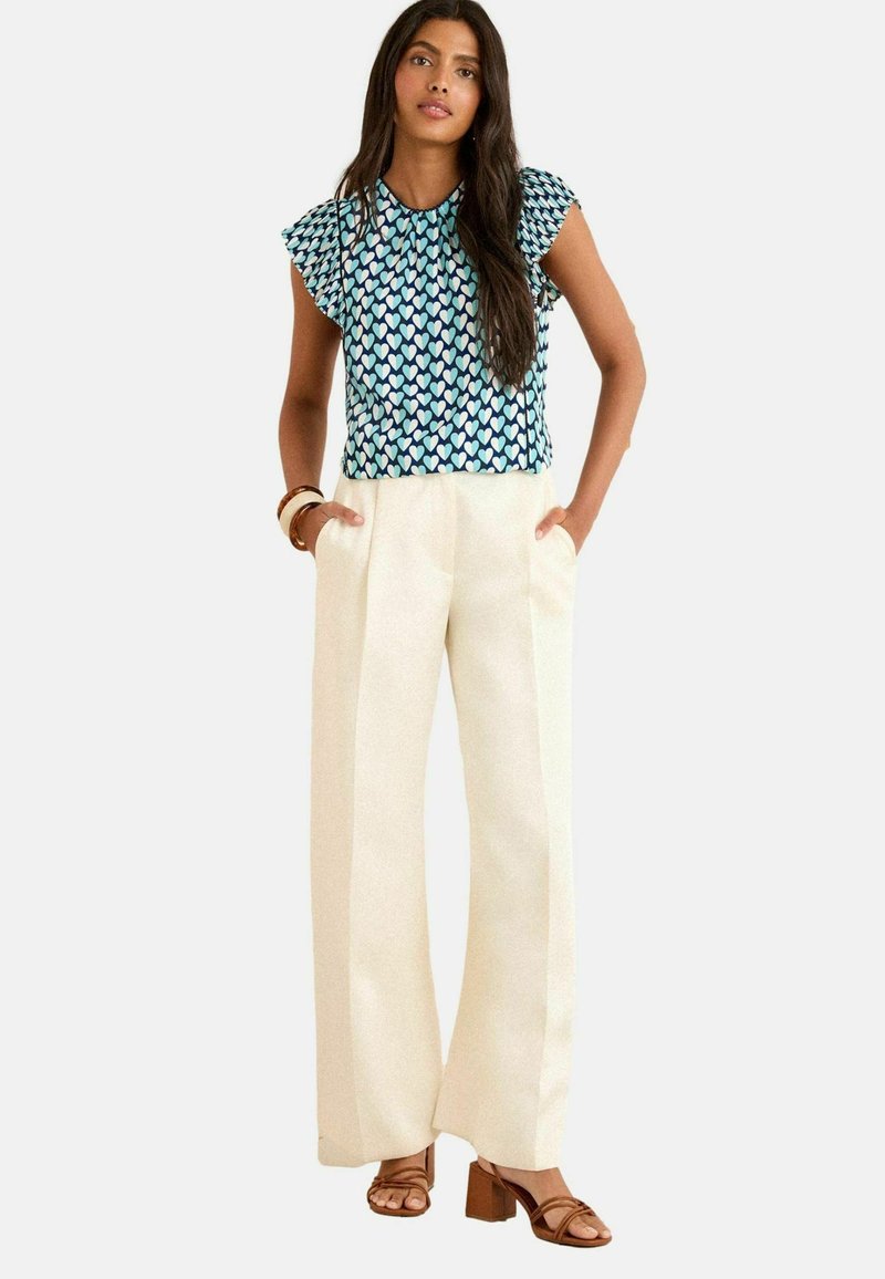 Blå och grönmönstrad topp med fladdrande ärmar, matchad med vita vida byxor. Modellen bär bruna klacksandaler med en slät yta.