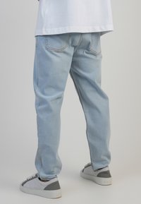 Jeans de mezclilla de color azul claro con un ajuste relajado, que cuentan con bolsillos traseros y detalles de costura. Combinados con zapatillas grises y una camiseta blanca.